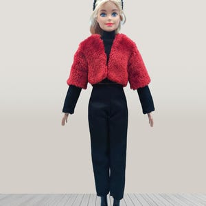 May include: A doll wearing a red faux fur cropped jacket, a black turtleneck, black pants, and black boots. The doll has blonde hair and is wearing a black headband. The doll is standing on a light wood-look surface.