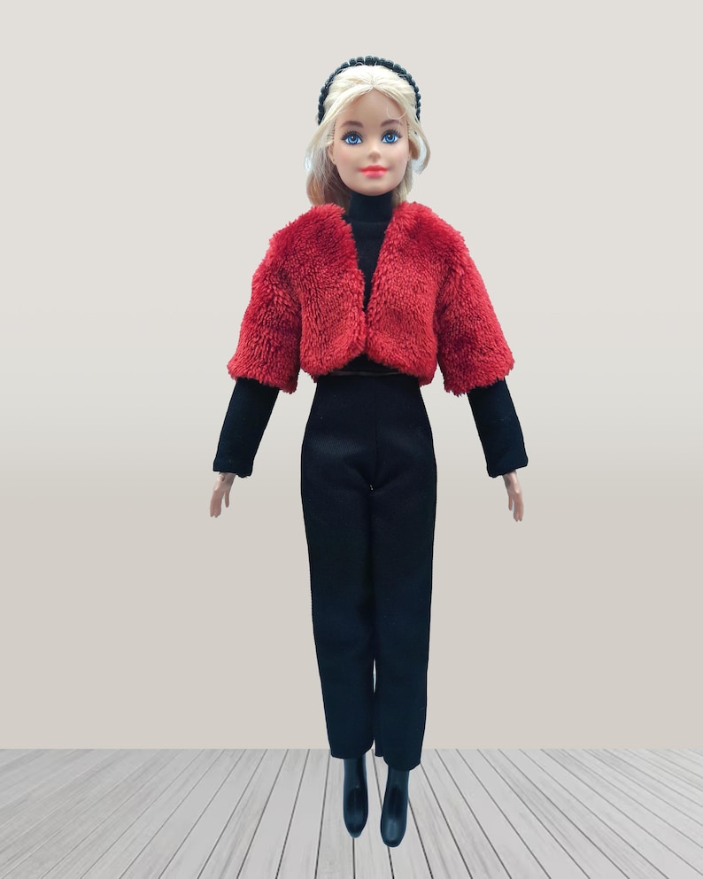 May include: A doll wearing a red faux fur cropped jacket, a black turtleneck, black pants, and black boots. The doll has blonde hair and is wearing a black headband. The doll is standing on a light wood-look surface.