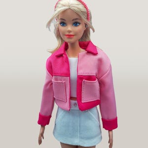 May include: A doll wearing a two-tone pink jacket with pockets, a white top, and a light blue denim skirt. The doll has blonde hair and a pink headband. The jacket has dark pink trim and cuffs. The doll is wearing a white belt.