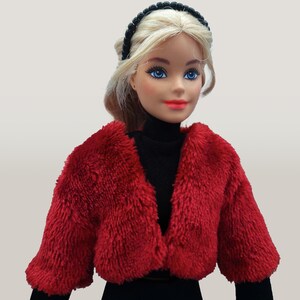 May include: A doll with blonde hair, blue eyes, and red lipstick, wearing a black turtleneck and a short, fluffy red jacket. A black beaded headband adorns the doll's hair. The doll is dressed in a fashion-forward style.