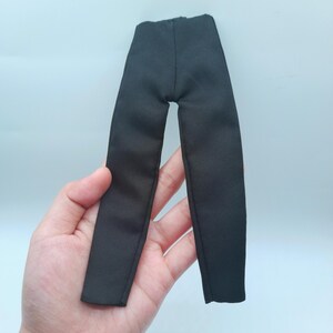 May include: Black doll-sized pants made of a textured fabric. The pants are a solid black color and appear to be tailored. The garment is designed for a doll and is held in a hand against a light blue background.