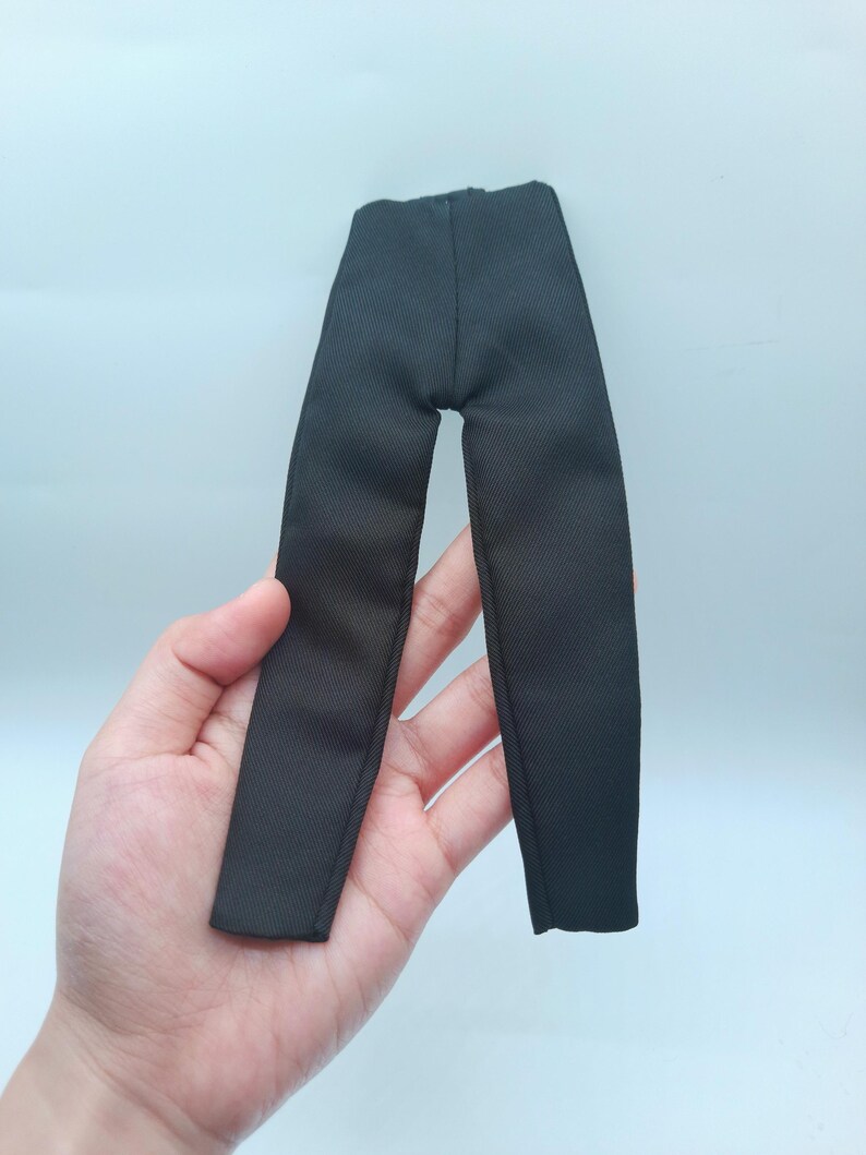 May include: Black doll-sized pants made of a textured fabric. The pants are a solid black color and appear to be tailored. The garment is designed for a doll and is held in a hand against a light blue background.
