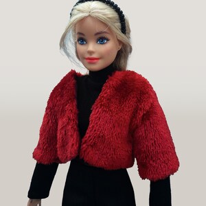 May include: A doll wearing a red faux fur bolero jacket over a black turtleneck and black pants. The doll has blonde hair, blue eyes, and red lipstick. A black beaded headband completes the look.