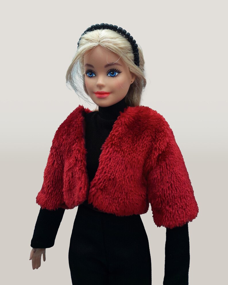 May include: A doll wearing a red faux fur bolero jacket over a black turtleneck and black pants. The doll has blonde hair, blue eyes, and red lipstick. A black beaded headband completes the look.