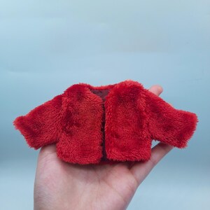 May include: A small, bright red plush jacket with a soft, fuzzy texture. The jacket has long sleeves and a simple, open front design. The garment is likely for a doll or small toy.
