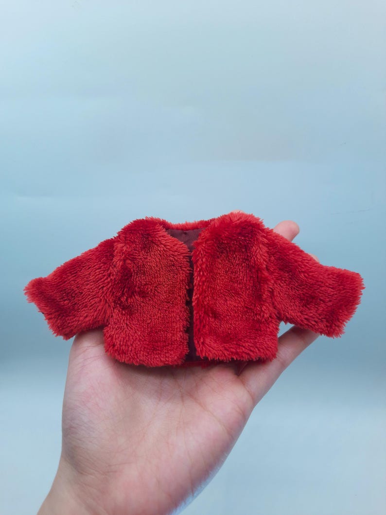 May include: A small, bright red plush jacket with a soft, fuzzy texture. The jacket has long sleeves and a simple, open front design. The garment is likely for a doll or small toy.