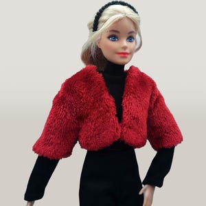 May include: A doll wearing a red faux fur bolero jacket over a black turtleneck and black pants. The doll has blonde hair, blue eyes, and a black headband. The outfit is set against a neutral background.