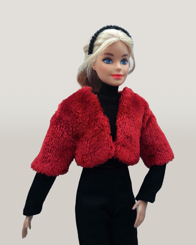 May include: A doll wearing a red faux fur bolero jacket over a black turtleneck and black pants. The doll has blonde hair, blue eyes, and a black headband. The outfit is set against a neutral background.