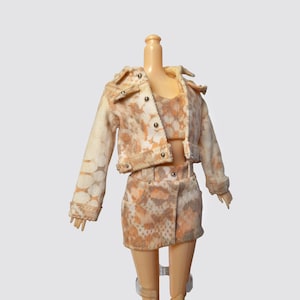 May include: A doll wearing a beige and white patterned denim jacket and matching skirt. The jacket has a collar and a button closure. The skirt is a mini skirt with a high waist.