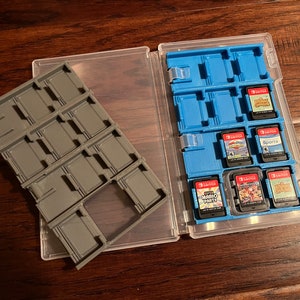 May include: A clear plastic case with a blue insert designed to hold video game cartridges. The case is open, revealing several game cartridges. A separate gray cartridge holder is next to the case. The games are labeled with various titles.