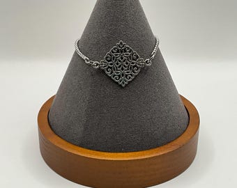 Könnte beinhalten: Ein silbernes Armband mit einem filigranen, komplizierten, rautenförmigen Design. Das Armband wird auf einem Holzständer präsentiert.