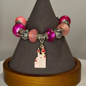 Op de afbeelding: Een roze en zilveren armband met een roze kasteelbedel. De armband heeft verschillende kralen, waaronder roze, wit en zilver.