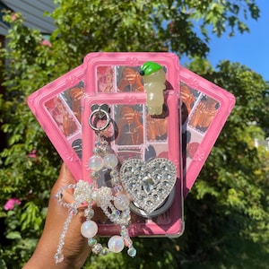 May include: Pink phone case with a framed photo design, a rhinestone heart pop socket, and a decorative beaded charm. The case also features a clear, green-hatted figurine.