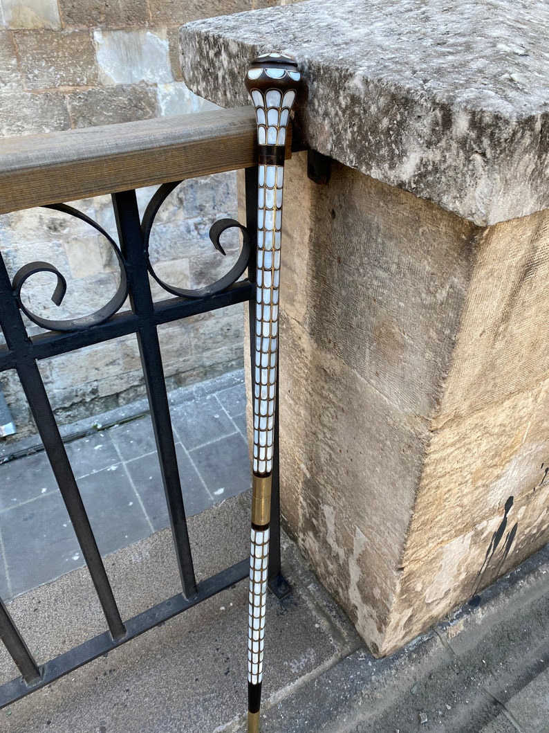 Puede incluir: Un bast&oacute;n blanco y dorado con una parte superior decorativa. El bast&oacute;n est&aacute; hecho de madera y tiene un patr&oacute;n blanco y dorado.