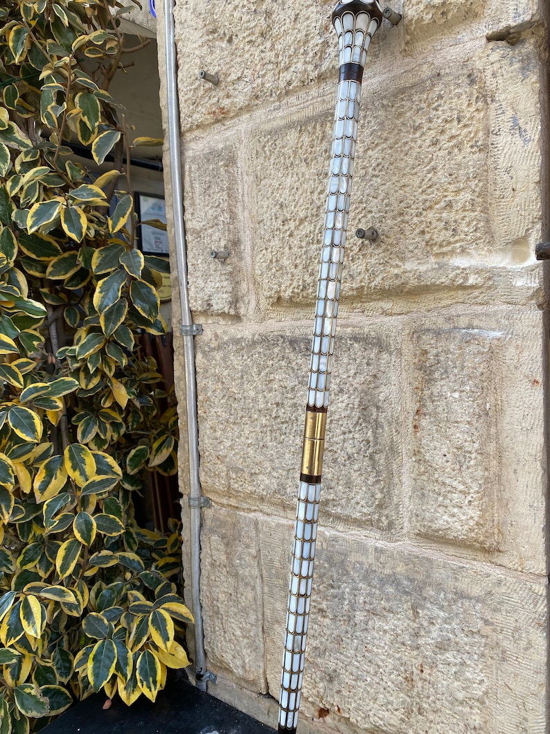 Puede incluir: Un bast&oacute;n blanco y dorado con una parte superior decorativa. El bast&oacute;n est&aacute; hecho de n&aacute;car y tiene una banda de lat&oacute;n alrededor del medio.