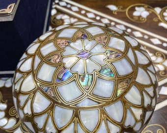 May include: A round, decorative box with a intricate design made of mother of pearl and gold. The box is on a wooden surface with a similar design.