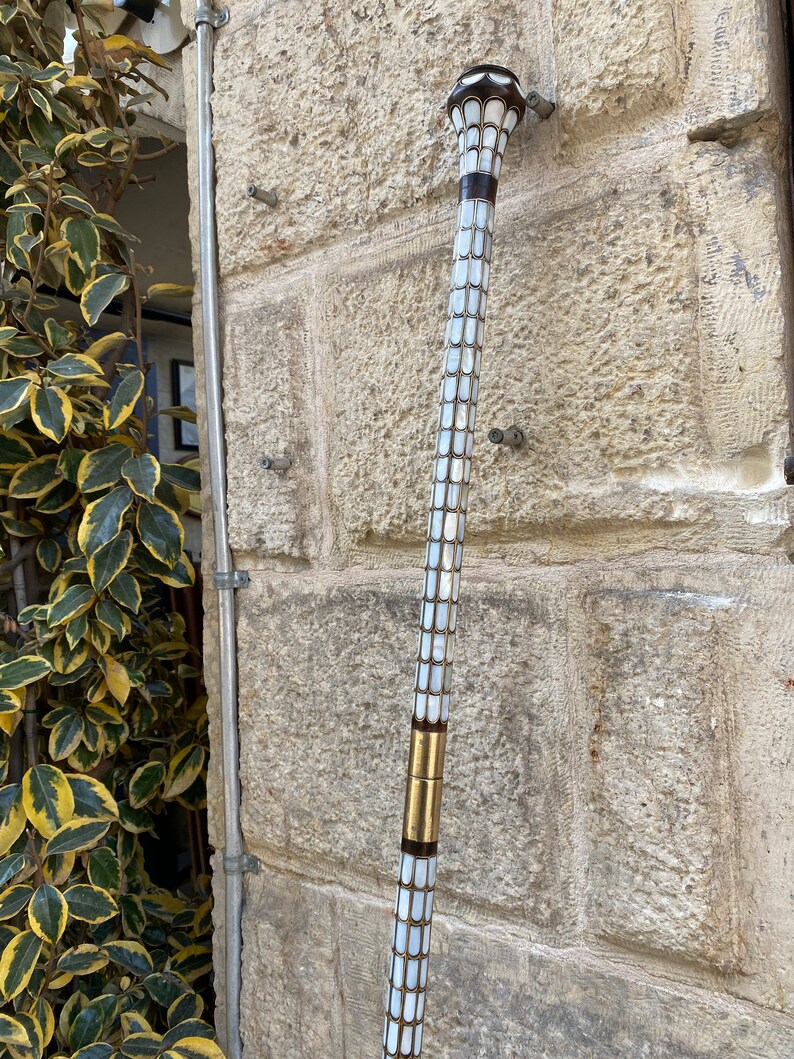 Puede incluir: Un bast&oacute;n de paseo con un eje de patr&oacute;n blanco y dorado y una parte superior de color marr&oacute;n oscuro. El bast&oacute;n est&aacute; hecho de madera y tiene un dise&ntilde;o decorativo.