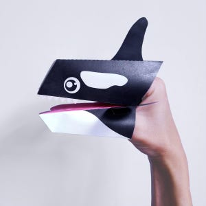Pode incluir: Fantoche de orca preto e branco com língua rosa e dentes brancos. O fantoche tem uma grande barbatana dorsal e um olho branco. O fantoche é feito de papel.
