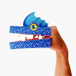 May include: Blue and pink paper puppet of a dragon with a large, open mouth and white teeth. The dragon has a blue fin on its head and a yellow eye.
