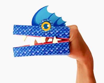Puede incluir: Marioneta de papel azul y rosa de un dragón con una boca grande abierta y dientes blancos. El dragón tiene una aleta azul en la cabeza y un ojo amarillo.
