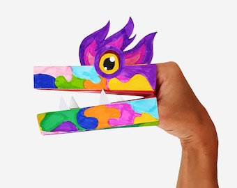 Puede incluir: Un títere de dedo de papel colorido con forma de monstruo con un ojo grande y dientes afilados. El títere está hecho de cartón y decorado con colores brillantes y patrones.