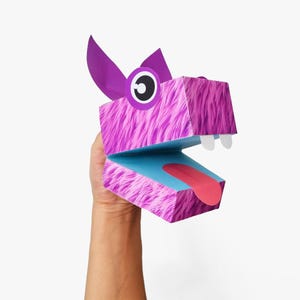 May include: A purple and blue paper puppet with a fuzzy texture and a large, open mouth. The puppet is held by a hand.