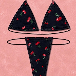 May include: A black two-piece bikini set featuring a repeating cherry print with red cherries and green leaves. The top consists of two triangular cups with thin black string ties, and the bottom is a thong-style piece with thin side ties.