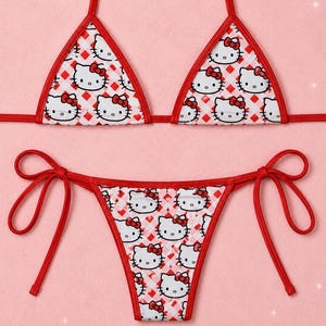Puede incluir: Un conjunto de bikini de dos piezas con base de tela blanca, un patrón repetitivo de diamantes rojos y caras del personaje Hello Kitty. La parte superior tiene copas triangulares con bordes rojos y lazos, la parte inferior tiene lazos laterales.
