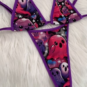 May include: A purple and black bikini set featuring a repeating pattern of cartoon ghosts, hearts, and stars. The bikini has purple trim and straps. The background of the pattern is a dark space-like design.
