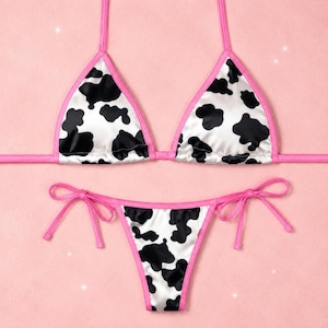 May include: A two-piece bikini set featuring a white fabric with a black cow print pattern. The top consists of two triangular cups with thin pink straps, and the bottom is a matching bikini brief with side tie closures and pink trim.