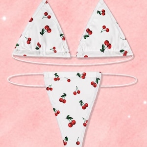 May include: A white two-piece bikini set featuring a repeating cherry print with red cherries and green leaves. The top consists of two triangular cups with thin white string ties, and the bottom is a high-cut thong style with thin side ties.