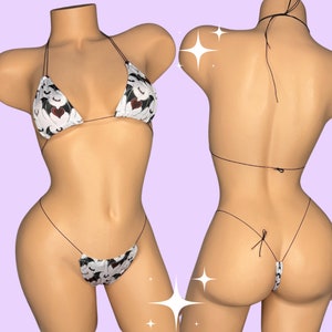 May include: A two-piece bikini set featuring a white fabric with a repeating pattern of black bats and red hearts. The top is a triangle style with thin straps, and the bottom is a thong style. The bikini is displayed on a mannequin.