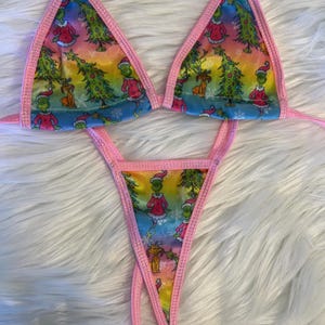 May include: A two-piece bikini set featuring a colorful print of a cartoon character and Christmas trees. The bikini top is a triangle style, and the bottoms are a thong style. The edges are trimmed with pink fabric.