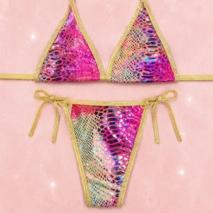 May include: A two-piece bikini set featuring a vibrant pink and iridescent snakeskin pattern. The triangle top and high-cut bottom are trimmed with metallic gold fabric and include thin gold side ties for an adjustable fit and secure closure.
