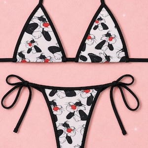 May include: A two-piece bikini set featuring a white fabric base with an all-over repeating print of the cartoon character Sylvester the Cat. The top consists of two triangular cups with black trim, while the bottom features side tie closures.