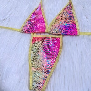 Puede incluir: Un conjunto de bikini de dos piezas con un vibrante estampado de piel de serpiente en tonos rosa, naranja y amarillo. La parte superior triangular y la braguita de estilo tanga están ribeteadas con tela dorada. El bikini se muestra sobre una superficie blanca y esponjosa.