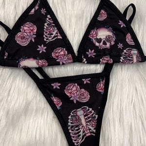 Puede incluir: Conjunto de bikini negro con estampado floral y de esqueleto. La parte superior es de estilo triangular y la parte inferior es un tanga.