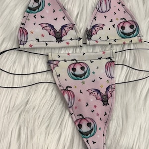 May include: A two-piece Halloween-themed lingerie set. The light pink fabric features a pattern of jack-o'-lanterns, bats, and stars. The top is a triangle bikini style, and the bottom is a thong. Black string ties are attached.