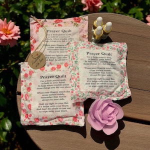May include: Three prayer quilts with floral patterns and text, a paper flower, and essential oil bottles. The quilts have a pink and white floral design with the words "Prayer Quilt". A tag reads "Handmade with love".