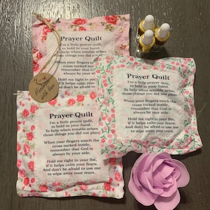 May include: Three prayer quilts with floral patterns and text, alongside small glass bottles with gold lids. A paper rose is also present. The quilts have a tag that says "Handmade with love."