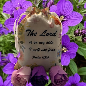 May include: A cream-colored drawstring bag with floral patterns and the text "The Lord is on my side, I will not fear, Psalm 118:6." The bag is surrounded by purple flowers and three artificial roses.