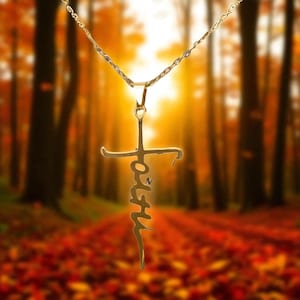 May include: A gold-colored pendant necklace featuring a cross design with the word "Faith" inscribed vertically. The necklace hangs against a blurred backdrop of a forest with autumn foliage.