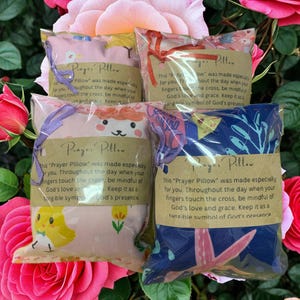 May include: Four small, packaged prayer pillows with floral and animal designs, tied with ribbons. Each pillow has a tag with text. The pillows are set against a backdrop of pink roses.