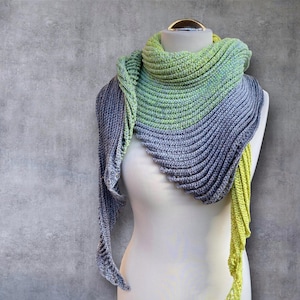 May include: A gray and green crocheted shawl with a fringed edge. The shawl is draped over a white mannequin.