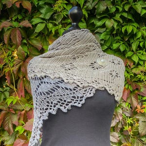 May include: A grey and white crocheted shawl with a delicate lace pattern. The shawl is draped over a dress form and has a decorative button.