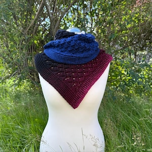 Puede incluir: Exhibición de tres artículos de punto: un cuello negro, un cuello azul y un chal burdeos. El chal tiene forma triangular con un patrón geométrico. Los artículos se exhiben en un maniquí blanco sobre un fondo de follaje verde.