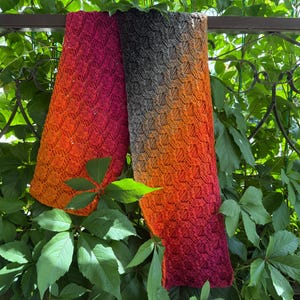May include: A knitted scarf with a gradient of colors, from deep red to orange and gray. The scarf is hanging against a backdrop of green leaves and a decorative metal structure. The scarf's texture is visible.