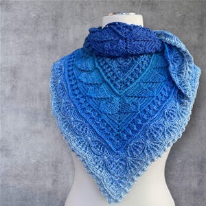 May include: A blue knitted shawl with a gradient effect, transitioning from deep blue at the top to a lighter shade at the bottom. The shawl features intricate patterns and a scalloped edge, displayed on a mannequin.