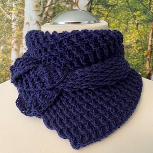 Può includere: Un collo lavorato a maglia blu scuro. La sciarpa è realizzata con un filato spesso e strutturato e presenta un design attorcigliato sul davanti. Il collo è esposto su un manichino, mostrando la sua forma e il suo stile. Lo sfondo è una vista sfocata di alberi.