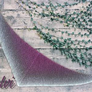 May include: A triangular crocheted shawl with a gradient of light gray to purple. The shawl is laid out on a wooden surface with green vines growing on the wall behind it. The word "Talvi" is written in cursive in the bottom left corner of the image.